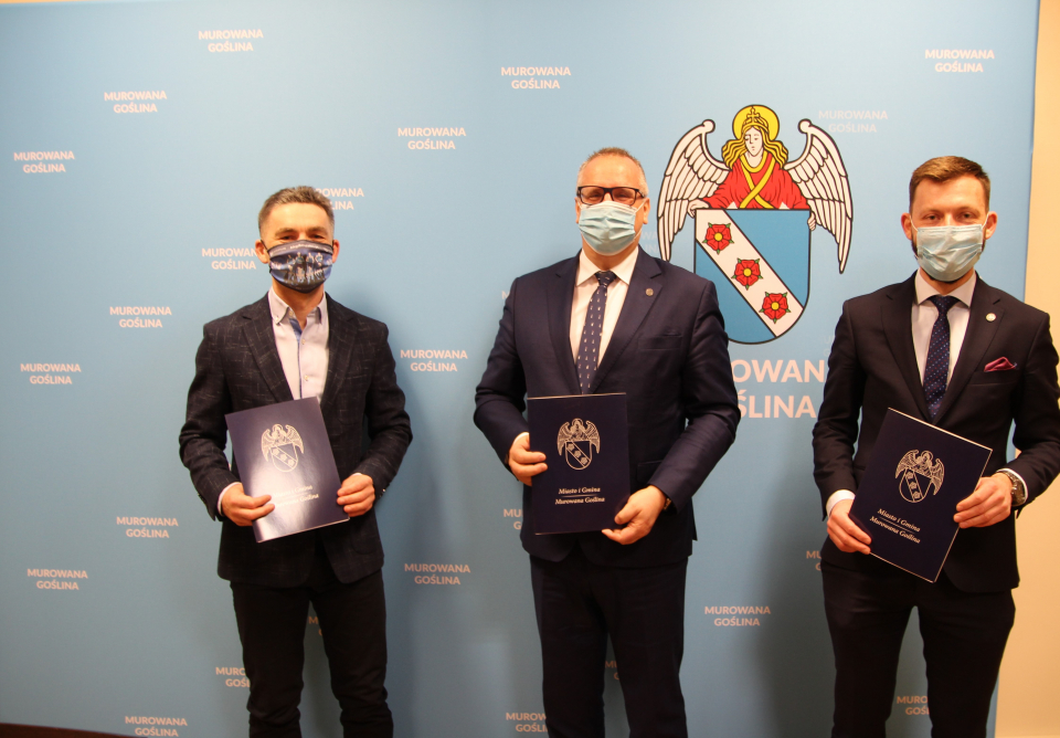 Zdjęcie - mężczyźni na tle planszy z napisami Murowana Goślina i herbem miasta