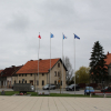 Zdjęcie - maszty z flagami, pomnik widoczny z daleka, pod nim wiązanki kwiatów i palący się znicz