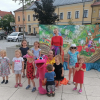 Dzieci pozują do wspólnego zdjęcia z zabawnym pluszowym ludzikiem i aktorką na tle dekoracji teatralnych.