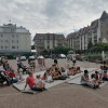 Publiczność siedząca na placu przy fontannie. W tle budynki i drzewa.