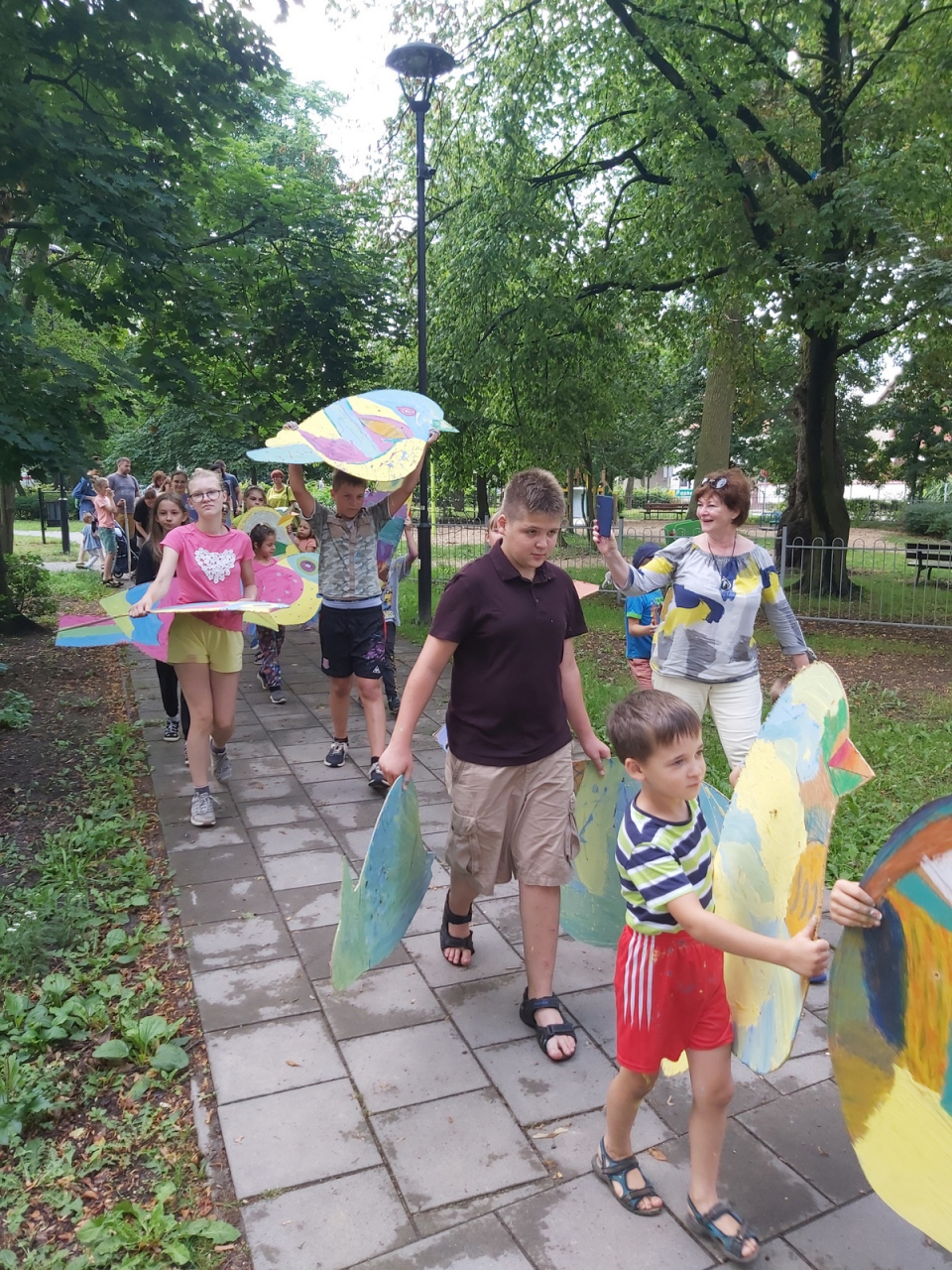 Przemarsz grupy dzieci z kolorowymi ptakami przez park.