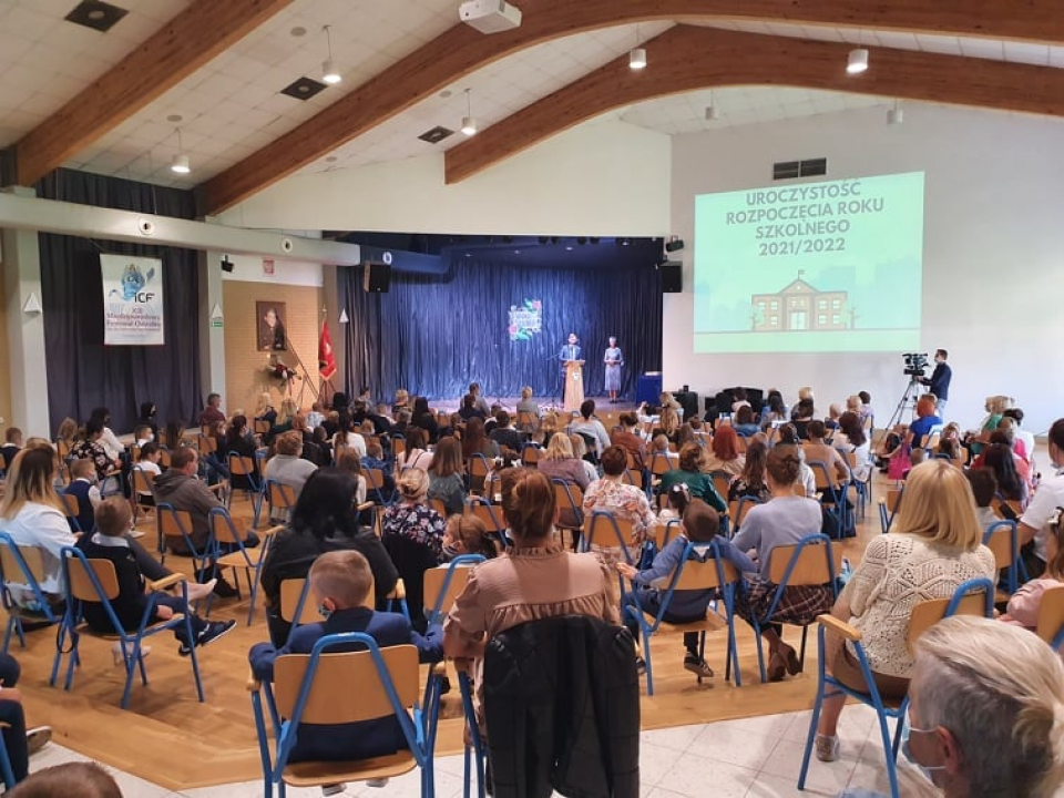 aula w szkole, przy mównicy burmistrz, na 1 planie nauczyciele i uczniowie, siedzą na krzesłach