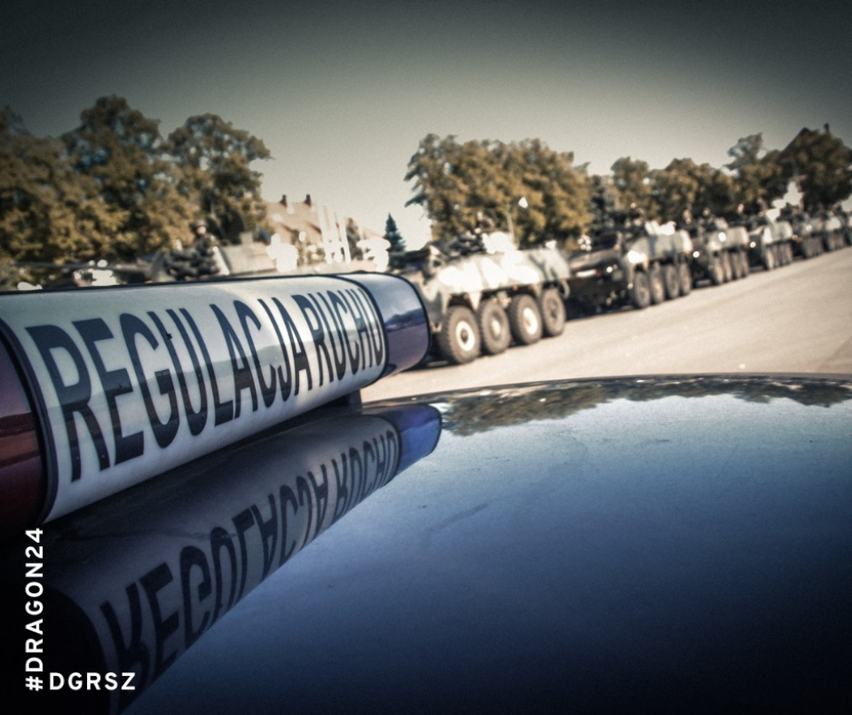 tzw. "kogut" na dachu pojazdu z napisem policja, w tle ciąg samochodów wojskowych