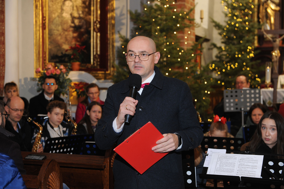 mężczyzna z mikrofonem, w tle orkiestra dętai choinka