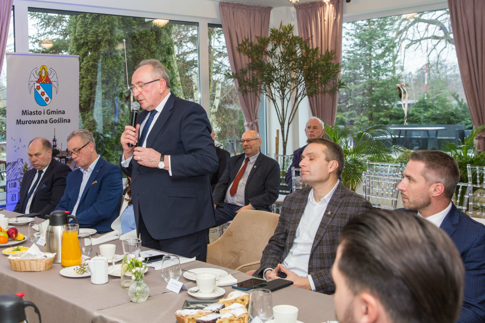 ludzie siedzą przy stole, mężczyzna stoi i przemawia do mikrofonu