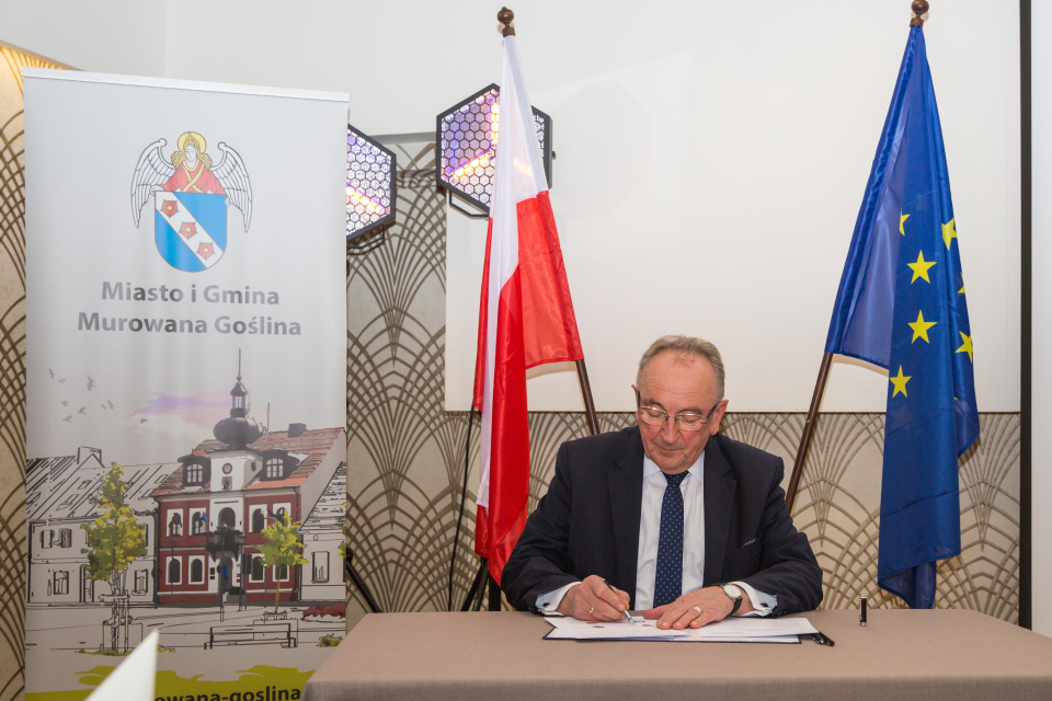 mężczyzna podpisuje dokument, w tle rollup z ratuszem i flagi RP i EU