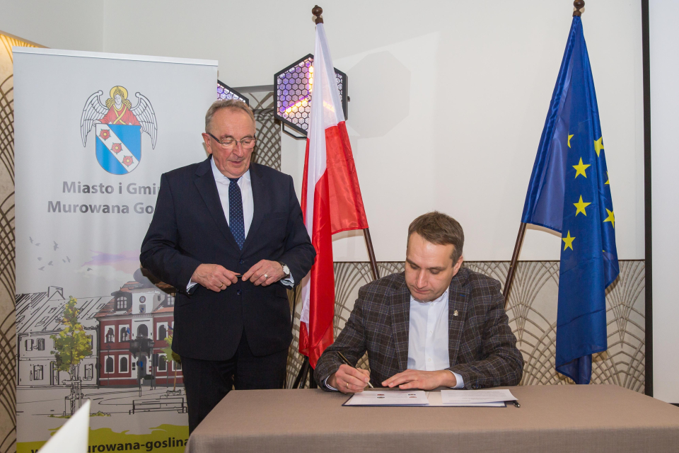 mężczyzna podpisuje dokument, obok mężczyzna, w tle rollup z ratuszem i flagi RP i EU