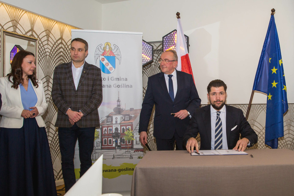 mężczyzna siedzi przy stoliku, obok 2 mężczyzn i kobieta, w tle rollup z ratuszem i flagi RP i EU