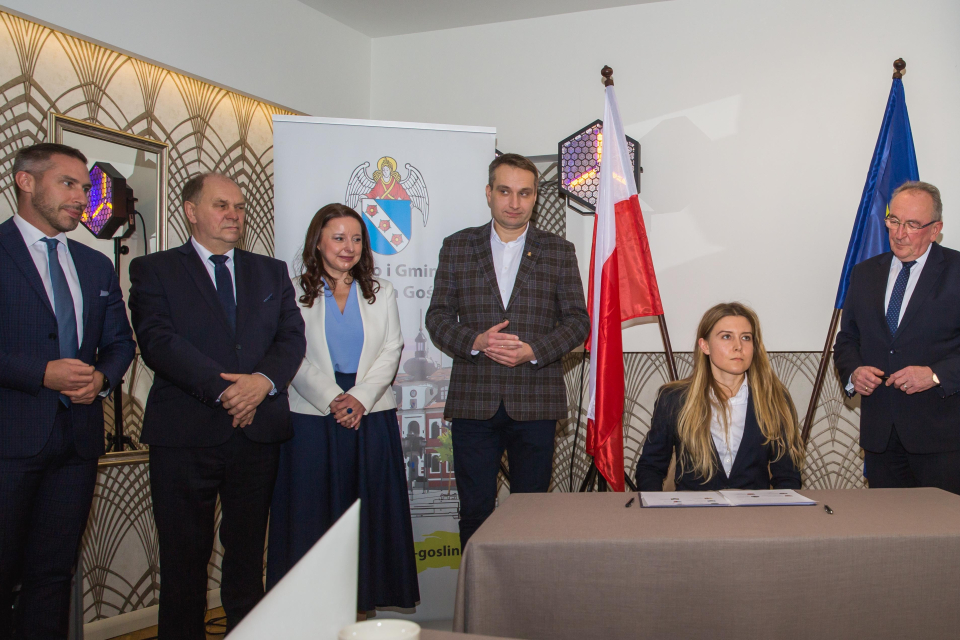 kobieta siedzi przy stoliku, obok 4 mężczyzn i kobieta, w tle rollup z ratuszem i flagi RP i EU