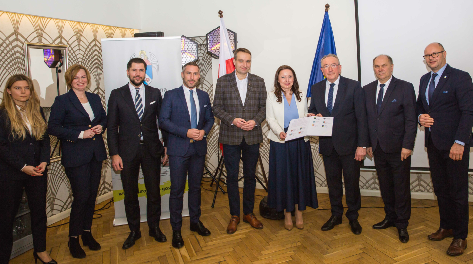 grupa 9 osób stoi na tle flag RP i UE, kobieta trzyma w rękach teczkę