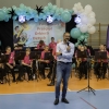 mężczyzna mówi do mikrofonu przed orkiestrą dętą, która siedzi na krzesłach na hali widowiskowo-sportowej