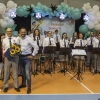 dyrygent z kwiatami i upominkiem w towarzystwie Organizatorów, w tle muzycy orkiestry dętej