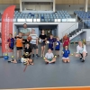 dzieci na sali gimnastycznej trzymają piłki, kobieta obok kuca trzymając hula hop, za dziećmi stoi mężczyzna