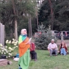 kobieta stoi na trawie, w tle mężczyzna grający na akordeonie oraz kobieta i mężczyzna na ławce