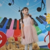 2. Na tle kolorowej scenografii stoi dziewczynka w różowej sukience stoi z mikrofonem w ręku.