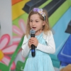 4. Na tle kolorowej scenografii stoi dziewczynka w niebieskiej sukience i z wiankiem na włosach ma mikrofon w rękach.