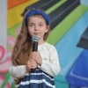 6. Na tle kolorowej scenografii stoi dziewczynka z długimi włosami i w opasce na głowie z mikrofonem w ręku.