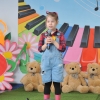 7. Na tle kolorowej scenografii stoi dziewczynka w koszuli, kaloszach z mikrofonem w ręku, przed nią stoi konewka.