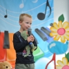 12. Na tle kolorowej scenografii, stoi chłopiec w ciemnej koszulce trzyma w ręku mikrofon i maskotkę.