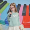 Na tle kolorowej scenografii stoi dziewczynka śpiewa do mikrofonu