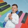 Na tle kolorowej scenografii stoi chłopczyk i śpiewa do mikrofonu