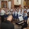 dyrygentka z chórem dziewczęcym przed ołtarzem, po boku kobieta akompaniująca na instrumencie klawiszowym