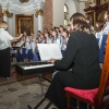 dyrygentka z chórem dziewczęcym przed ołtarzem, po boku kobieta akompaniująca na instrumencie klawiszowym