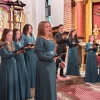 Kobieta w długiej zielonej sukience stoi i śpiewa, za nią w tle widać innych chórzystów.