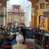 Chór stoi przed ołtarzem głównym, po bokach widać siedzącą publiczność w ławkach