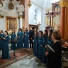 Jedna z chórzystek stoi i trzyma mikrofon, pozostali członkowie chóru w raz z dyrygentka stoją za nią.