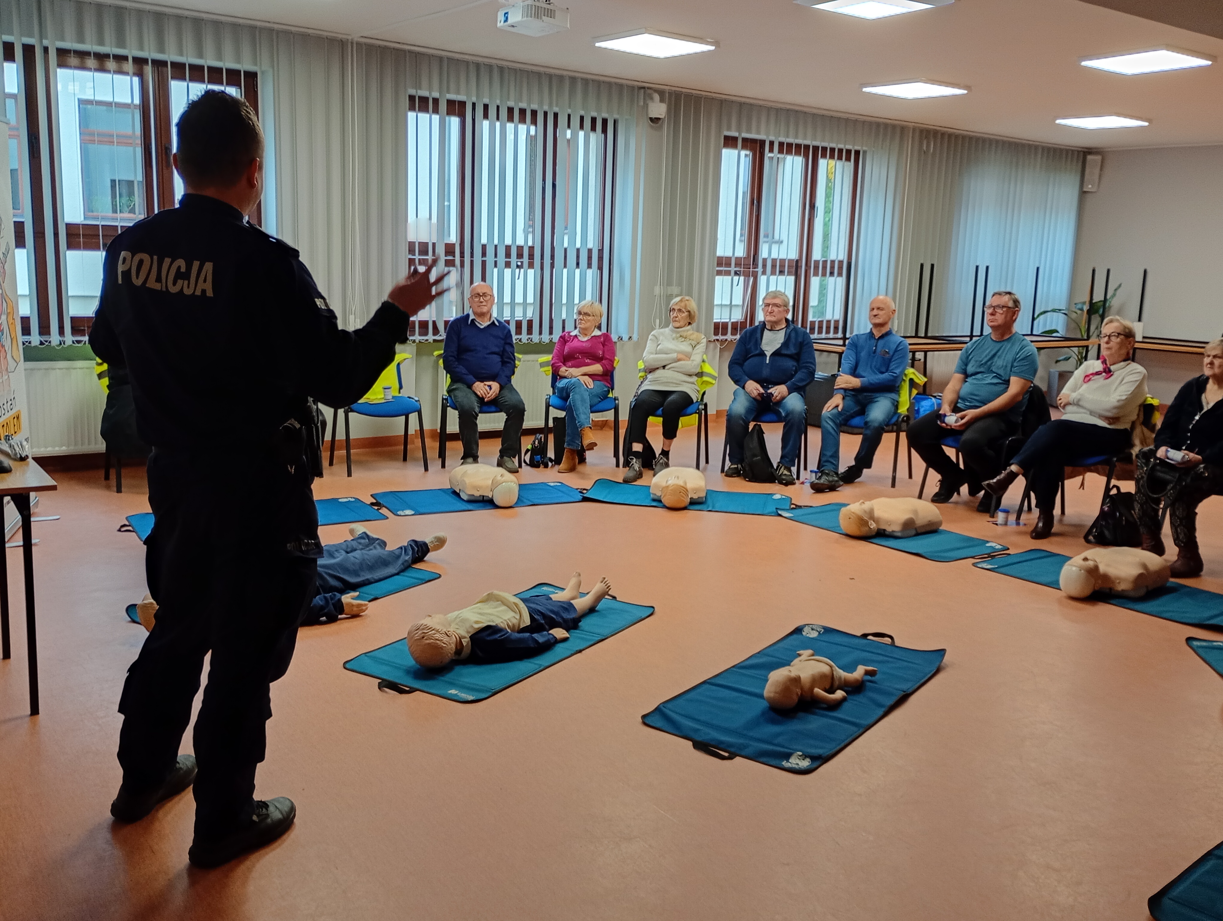 [obraz] policjant mówi do siedzących na sali seniorów