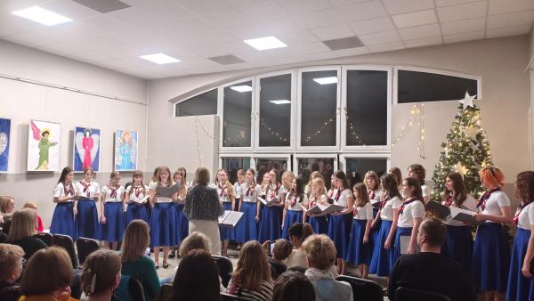 [obraz] Dziewczęcy Chór Canzona w niebieskich spódnicach stoi przed dyrygentką.