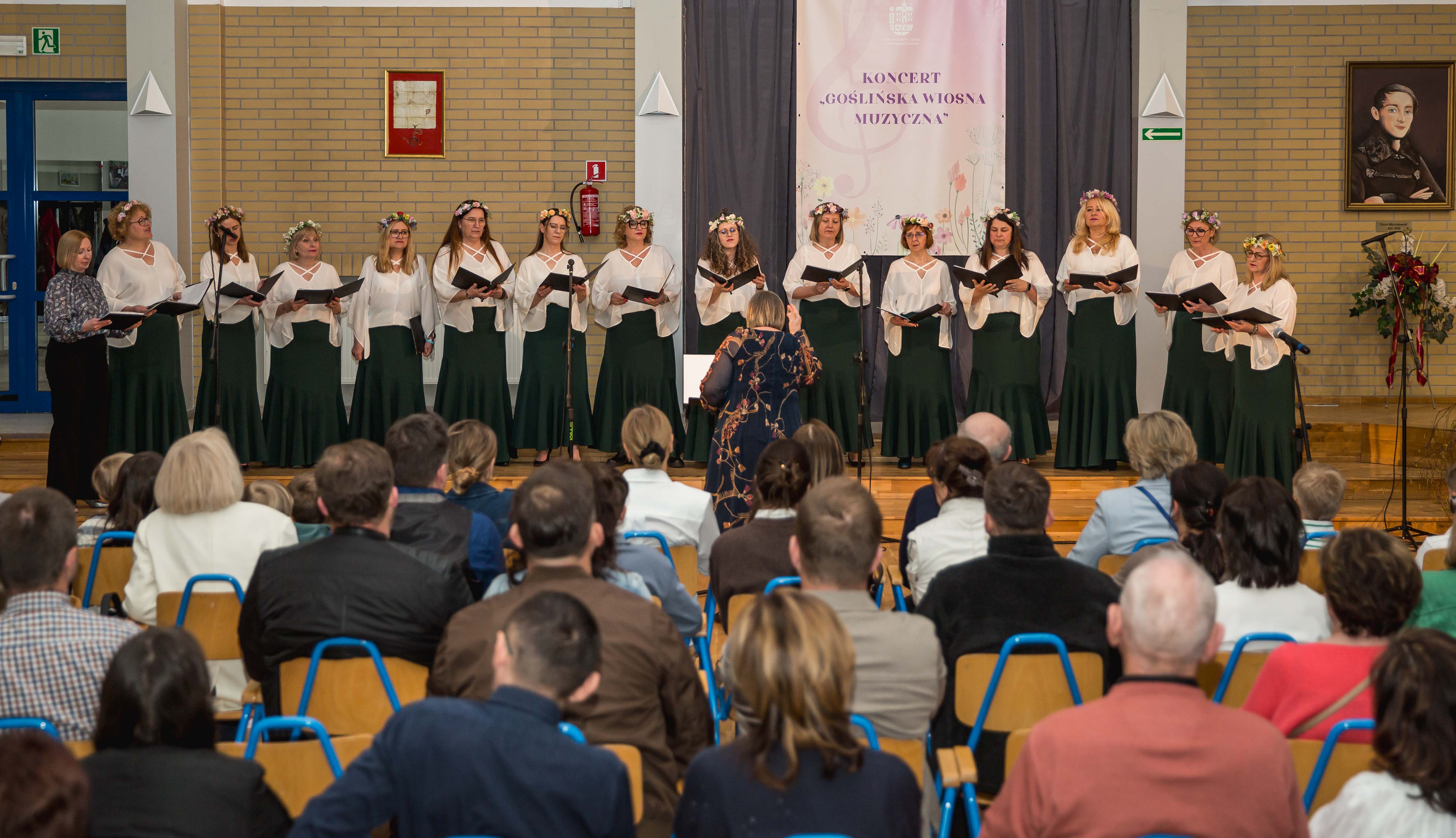[obraz] Na pierwszym planie publiczność, w tle Żeński Chór Canzona w białych bluzkach i zielonych spódnicach. Przed nimi stoi dyrygent Elżbieta Wtorkowska.